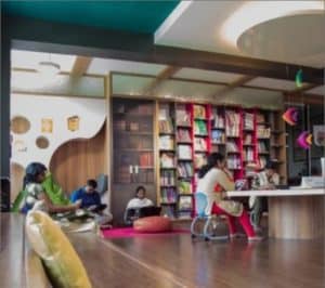 Inside view of School library with books arranged on the shelves and three staff sitting on the table and three more sitting down and reading