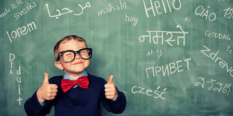 small boy wearing school uniform and spectacles standing in front of the school board smiling showing two hands indicating his passion for learning various languages