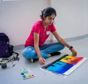 A girl student is making a color painting in a board, some water colors and bag can be seen beside her.