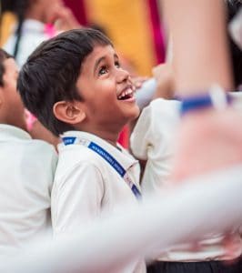 A small school boy with a school uniform and identify card on his neck is having a broad smile on his face.