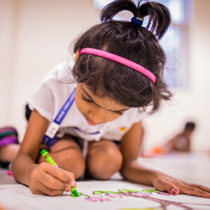 A small girl student is sitting on the floor and drawing something on a paper with the hA small girl student is sitting on the floor and drawing something on a paper with the help of pen and sketch.elp of pen and sketch.