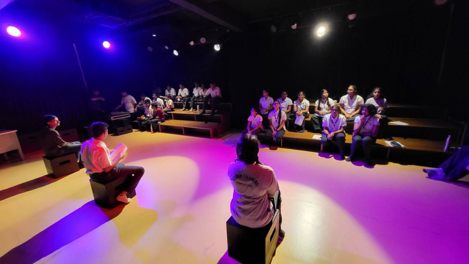 A set of students are watching three of their classmates involved in performing arts at the stage.